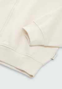 Veste zippée beige clair avec des poignets et un ourlet côtelés. Tissu doux avec une texture lisse. Présente une petite étiquette de marque sur le côté.