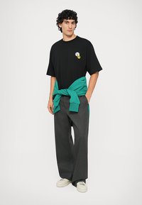 Μαύρο oversized μπλουζάκι με γραφικό μπροστά και κίτρινο χαρούμενο πρόσωπο, συνδυασμένο με γκρίζες φαρδιές παντελόνες και ένα πράσινο πουλόβερ δεμένο στη μέση.