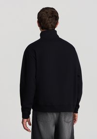 Felpa nera a collo alto con maniche lunghe, vestibilità rilassata e polsini a costine. Indossata con jeans in denim grigi, mostrando un design semplice.