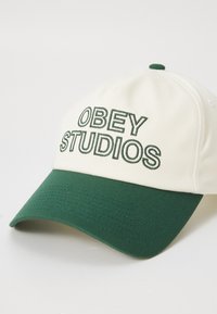 Căciulă de baseball cu un corp din material crem și o margine verde. Prezintă text brodat "OBEY STUDIOS" în verde. Suprafață texturată pe margine.