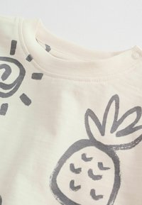 Svetlo kremna majica s sivimi motivi ananasa in sonca. Ima zaobljen izrez in pritrdilni gumb na rami. Mehka, teksturirana tkanina.