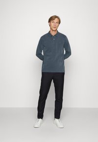 Cămașă polo cu mânecă lungă, de culoare albastră închisă, cu guler și închidere cu nasturi, asortată cu pantaloni negri și adidași albi; textură netedă.