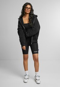 Črn zip-up hoodie prekrit s črnim majico s kratkimi rokavi in črnimi kolesarskimi hlačami z belim napisom "PEOUS". Bele superge z dodatki dopolnjujejo outfit.