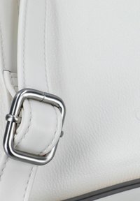 Esquina de un bolso de cuero blanco con una textura suave. Presenta un broche y una correa metálicos plateados, destacando los detalles de diseño minimalista.
