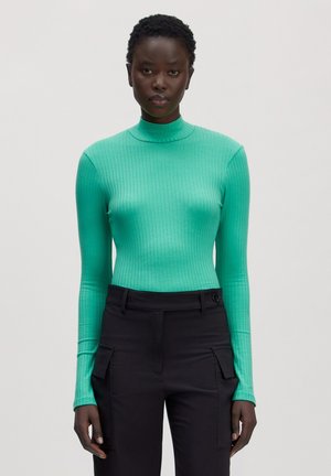 Frau trägt einen taillierten türkisfarbenen, gerippten Rollkragenpullover und hoch taillierte schwarze Cargohosen, steht vor einem schlichten hellen Hintergrund.