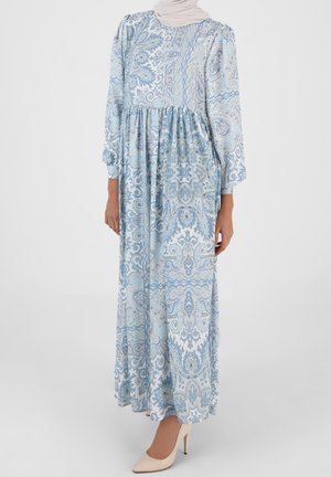 Femme portant une robe longue à manches longues bleu et blanche avec une taille froncée et des talons beiges.
