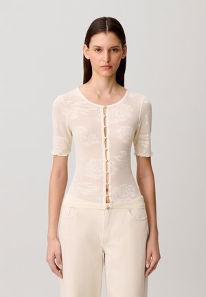 Mujer con una blusa de manga corta transparente color crema con estampado floral y botones, y pantalones de color claro, de pie frente a un fondo liso.