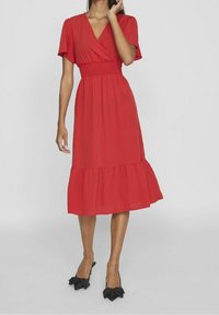 Červené midi šaty s překládaným designem, krátkými rukávy, nařaseným pasem a volánkovým lemem. Doplňují je černé lodičky s špičatou špičkou a mašlemi.