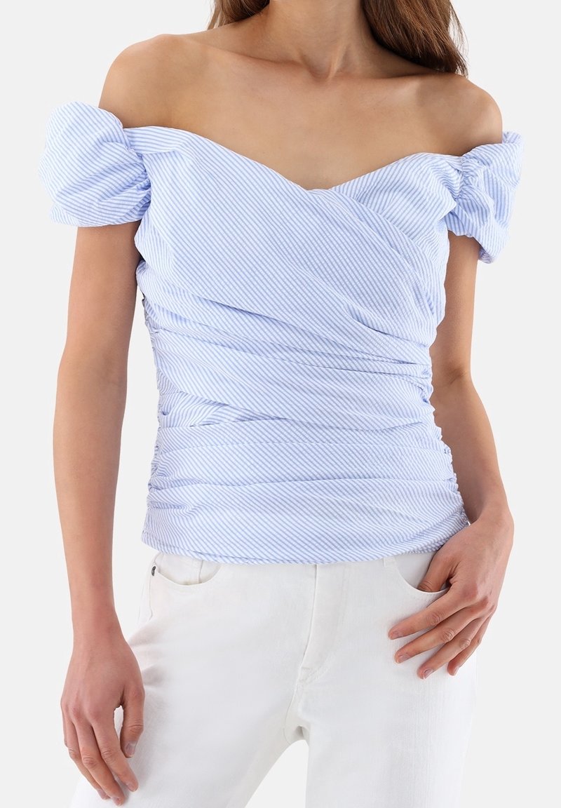 Femme portant un haut rayé bleu clair et blanc épaules dénudées avec des manches bouffantes et un pantalon blanc, main dans la poche.