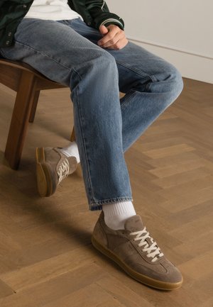 Bruine suède sneakers met witte veters, gumrubberen zool, gecombineerd met lichtblauwe spijkerbroeken en witte sokken, zittend op een houten stoel.