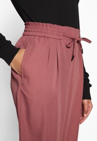 Lichtroze elastische taillebroek met een trekkoord. De stof is soepel met subtiele plooien en zijzakken.