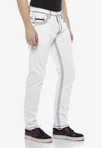 Weiße Jeans mit kontrastierenden schwarzen Nähten, schlanker Schnitt, fünf Taschen Design und mit Reißverschluss. Kombiniert mit schwarzen Sneakers.