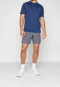Camiseta atlética azul de mangas cortas con cuello redondo, combinada con pantalones cortos grises y zapatillas de correr blancas. Presenta logo en la camiseta y los pantalones cortos.