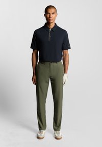 Mörkblå kortärmad polo med krage och gyllene detaljer, tillsammans med olivgröna byxor och ljusfärgade golfskoj. Vit handske på vänster hand.