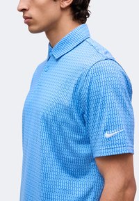 Άνδρας που φαίνεται από το πλάι, φοράει ένα γαλάζιο πουκάμισο πόλο Nike με σχέδια και λευκό λογότυπο Nike στο μανίκι.