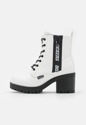 Bottines blanches avec un haut talon bloc noir, dotées de lacets noirs, d'une fermeture éclair et d'une bande logo noire sur le côté.