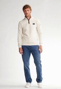 Pulover bej cu fermoar până la piept, manșete și tiv striate; material din amestec de bumbac cu un logo mic pe piept. Purtat cu blugi albaștri.