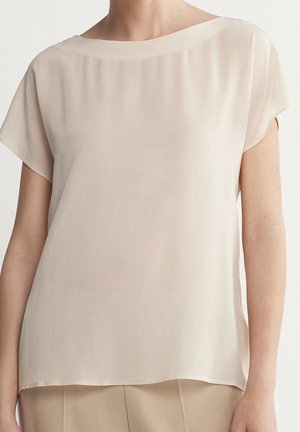 Femme portant un chemisier beige ample à manches courtes avec un col rond, assorti à un pantalon beige, sur un fond uni.