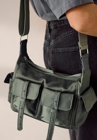 Zielona nylonowa torba na ramię z główną komorą zamykaną na zamek, dwoma przednimi kieszeniami na suwak i regulowanym paskiem. Gładka tekstura z kontrastującym przeszyciem.