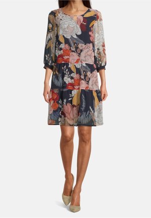 Femme portant une robe fleurie mi-longue à manches longues dans des tons bleu marine, rouge, rose, jaune et blanc, associée à des escarpins beige à bout pointu.