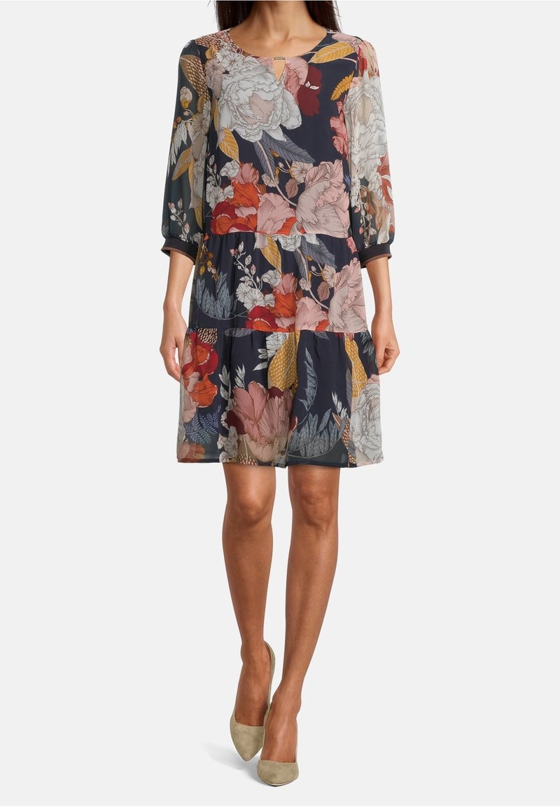 Femme portant une robe fleurie mi-longue à manches longues dans des tons bleu marine, rouge, rose, jaune et blanc, associée à des escarpins beige à bout pointu.