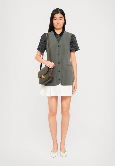 Jeune femme portant une robe sans manches gris à rayures, en dessous d'un polo noir à rayures et d'une jupe blanche, tenant un sac à bandoulière marron et portant des chaussures blanches à bouts pointus.