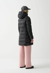 Schwarze Pufferjacke mit Kapuze, mit einem gesteppten Design und einem Logo-Akzent. Kombiniert mit hellrosa weiten Hosen.