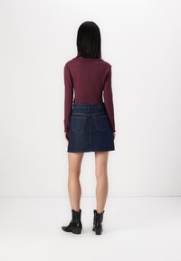 Burgundska rebrasta majica z dolgimi rokavi v kombinaciji s temno jeans kratko krilo. Krilo ima zadnje žepe in teksturiran videz. Črne gležnjarje.