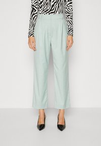  Lichtgroene wijde broek met een gestructureerde afwerking en plooien aan de voorkant, gecombineerd met zwarte puntige hakken. Gestreept langwerpig shirt zichtbaar.