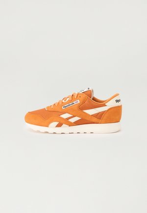 Baskets Reebok orange avec des bandes blanches, un lacet et une semelle blanche, vues de profil sur fond uni.