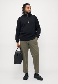 Svart zip-neck genser, olivengrønne chinos og svarte sko. Bærer en svart veske med teksturert detaljer. Enkel, uformell design.
