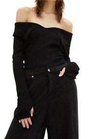 Femme portant un pull noir côtelé à épaules dénudées et un pantalon noir taille haute, une main dans la poche et l'autre posée le long du corps.
