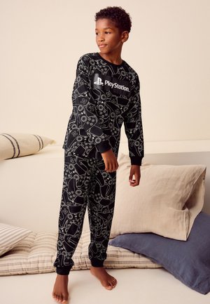 Dreng stående barfodet på puder iført sorte PlayStation-tema pyjamas med hvide controller-mønstre og logo på toppen.