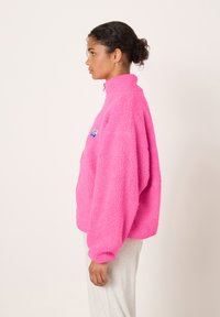 Roza pulover iz mehkega flisa z visokim ovratnikom, dolgimi rokavi in majhnim izvezenim logotipom na prsih. Mehka tekstura in sproščen kroj.