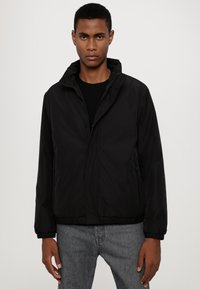 Giacca nera con zip e cappuccio, dotata di polsini e orlo elasticizzati, realizzata in tessuto morbido. Modello che indossa jeans grigi si trova contro uno sfondo neutro.