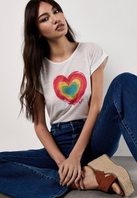 T-shirt blanc à manches courtes avec un motif de cœur multicolore. Associé à un jean flare bleu marine et des sandales espadrilles marron.