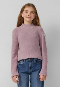 Meisje met lang haar in een geribbelde lavendelkleurige trui en blauwe jeans, staand tegen een effen lichtgrijze achtergrond.