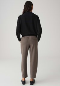 Schwarzer, gerippter Pullover mit hohem Kragen, kombiniert mit maßgeschneiderten grauen Hosen mit einer Gesäßtasche. Dazu schwarze Loafers.