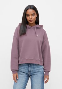 Paarse cropped hoodie met een trekkoordcapuchon, geribbelde manchetten en een klein boomlogo op de borst. Gecombineerd met blauwe jeans.