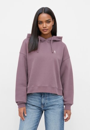 Lila gekürzter Hoodie mit einer Kordelzugkapuze, gerippten Bündchen und einem kleinen Baum-Logo auf der Brust. Kombiniert mit blauen Jeans.