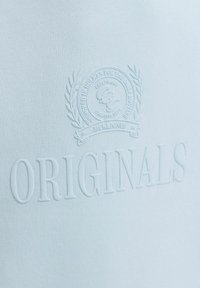 Tissu blanc texturé avec texte en relief "ORIGINALS" et un emblème circulaire au-dessus, comportant des branches de laurier et de petits détails textuels.