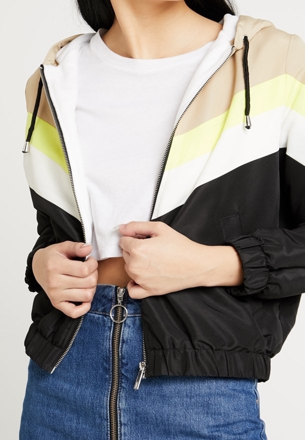 Chaqueta rompevientos colorida en negro, beige, blanco y franjas verde neón. Cuenta con capucha, cierre de cremallera y puños elásticos con bolsillos.