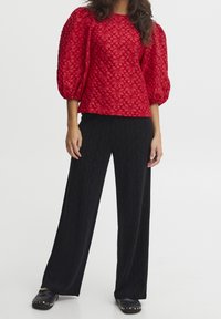 Haut rouge texturé avec des manches bouffantes, motif floral, associé à un pantalon large noir. Chaussures noires à enfiler avec des studs dorés.
