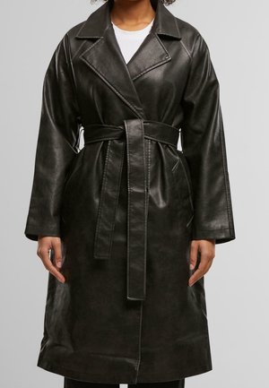 Woman wearing a black faux leather belted trench coat over a white top, standing against a plain light gray background.