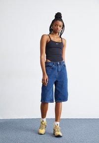 Top de alças preto justo com o logo da Nike, combinado com calções de ganga azuis até ao joelho. Usado com ténis amarelos e meias brancas até ao tornozelo.