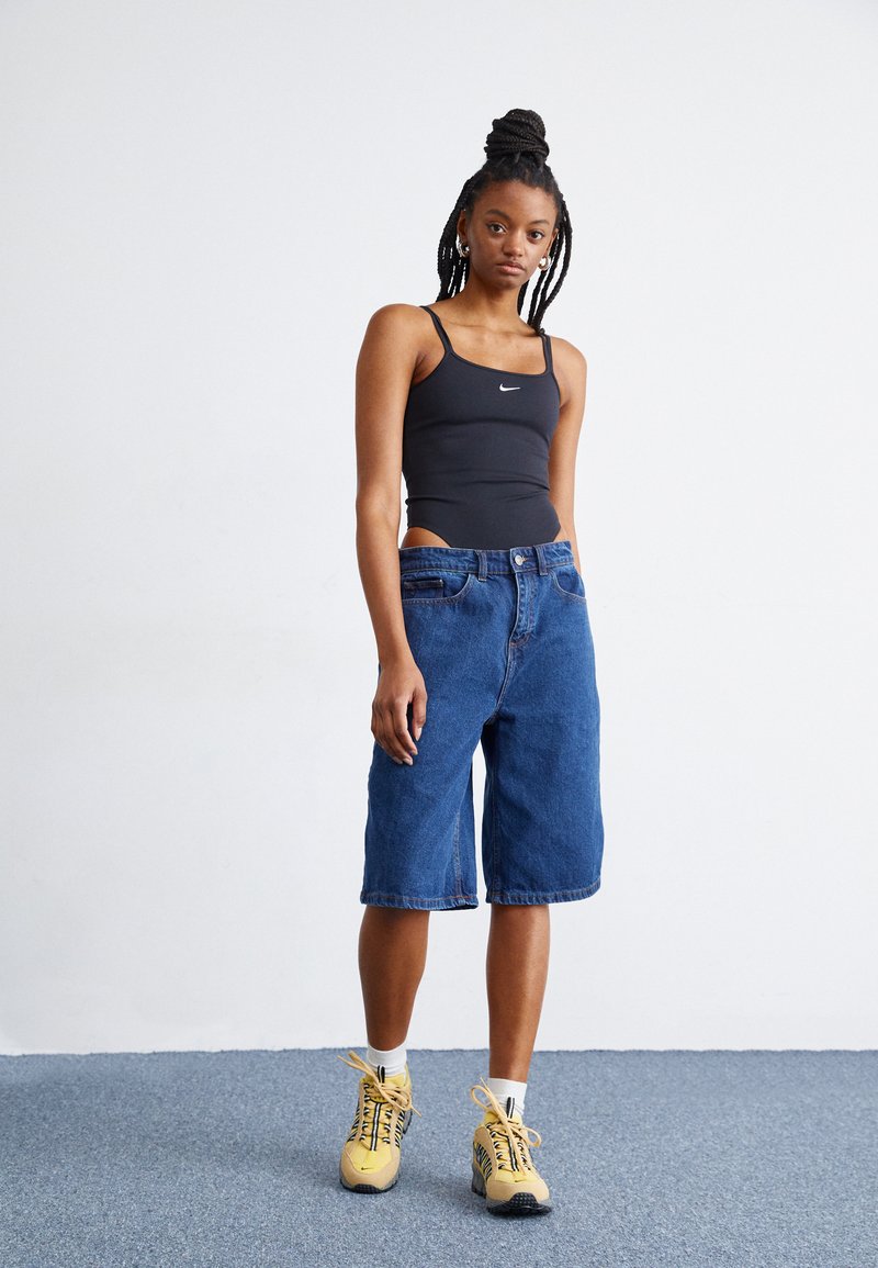 Débardeur noir ajusté avec un logo Nike, associé à un short en denim bleu arrivant aux genoux. Porté avec des baskets jaunes et des chaussettes blanches à la cheville.