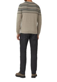 Man in een beige gebreide trui, zwarte jeans en bruine schoenen, staand met zijn rug naar de camera op een witte achtergrond.