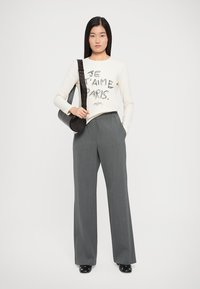 Lange grijze broek en een crèmekleurig shirt met lange mouwen en zwarte bedrukte tekst. Een zwarte crossbody tas met een ronde vorm. Zwarte schoenen.