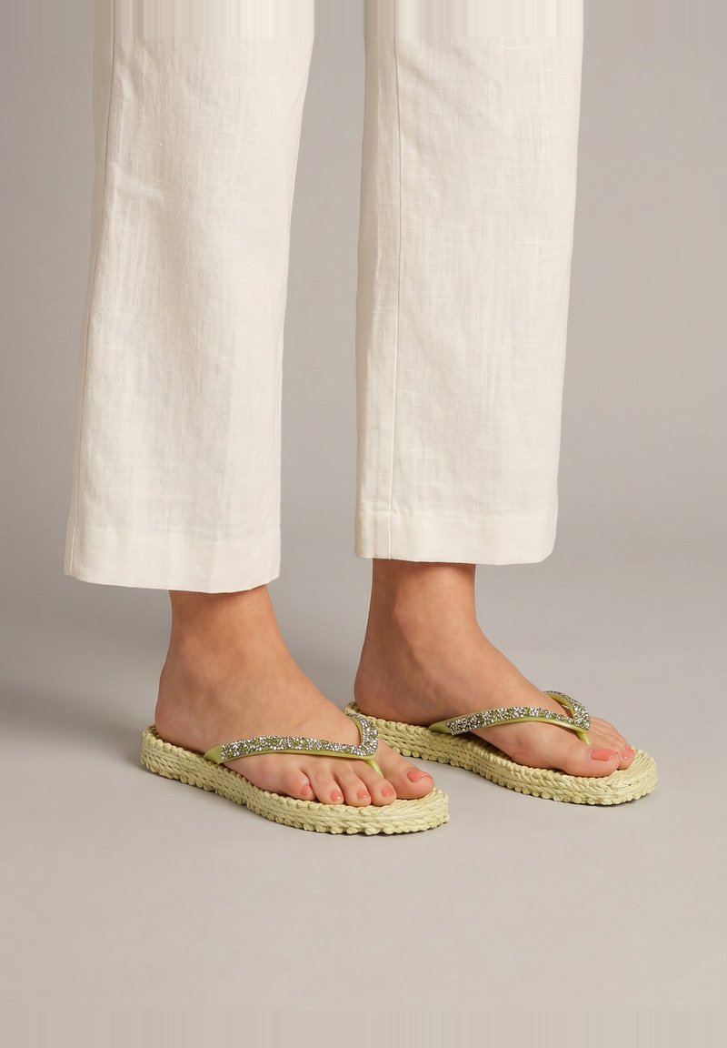 Pieds portant des tongs tressées beige avec des brides en strass, associées à un pantalon en lin blanc court sur un fond neutre.