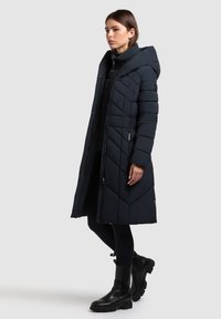 Vrouw staand in een zwarte lange doorgestikte jas met capuchon, zwarte leggings en zwarte enkellaarsjes tegen een effen lichte achtergrond.